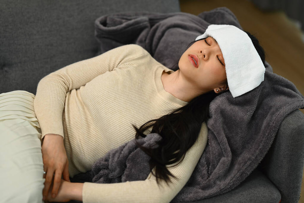 Хвора молода жінка лежить на дивані з мокрим рушником на лобі для зменшення високої температури. Концепція хвороби та охорони здоров'я. - Фото, зображення
