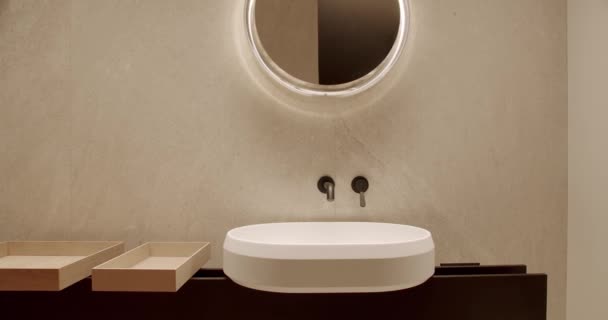 Μπάνιο με μοντέρνο εσωτερικό, κεραμικό washbowl, κρυφή βρύση, ξύλινο κουτί αποθήκευσης και στρογγυλό καθρέφτη κρέμεται στον τοίχο - Πλάνα, βίντεο