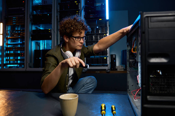 Молодой ИТ-техник, использующий фонарик, чтобы найти проблему с компьютером в современном дата-центре. Инженер за работой - Фото, изображение