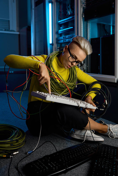Jovem mulher Técnico de TI envolto em fio de reparação teclado sentado no chão da sala do servidor - Foto, Imagem
