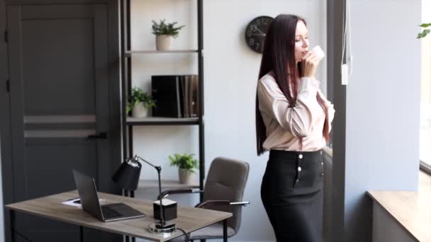 Όμορφη μελαχρινή γυναίκα που πίνει πρωινό καφέ, στέκεται κοντά στο γραφείο της στο γραφείο. Νεαρή γυναίκα που πίνει καφέ στο χώρο εργασίας - Πλάνα, βίντεο