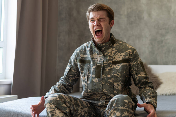 Gestresster Soldat, der in depressive Gedanken und Erinnerungen verstrickt ist. Traurig deprimierter junger Soldat in Tarnuniform sitzt auf der Couch und denkt an den Krieg. PTBS und Therapiekonzept - Foto, Bild