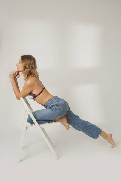 Fashion portrait of young woman in lace bra and wide leg blue jeans posing on the wooden chair on the white background - Photo, Image