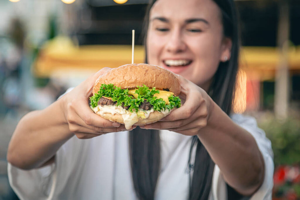 Egy vonzó fiatal nő kezében egy hamburgert tart, gyorskaját eszik az utcai kávézóban, homályos háttérrel.. - Fotó, kép