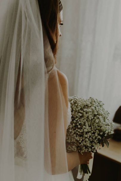 Porträt einer schönen jungen kaukasischen erkennbaren brünetten Braut, die tagsüber einen Strauß weißer Boutonnieres im Raum hält, Ansicht von unten, Nahaufnahme mit selektivem Fokus. - Foto, Bild