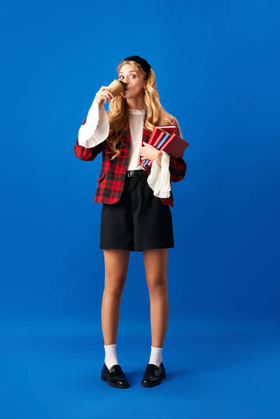 Portrait of young, female student dressed in university uniform standing holding books and drinking coffee against blue studio background. Concept of education, studying, youth, student lifestyle. Ad - Photo, image