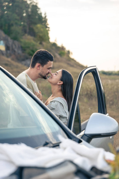 Ένας άντρας και μια γυναίκα φιλιούνται στο χωράφι, στέκονται δίπλα στο αυτοκίνητο, η έννοια των στενών σχέσεων και των διακοπών μαζί. - Φωτογραφία, εικόνα