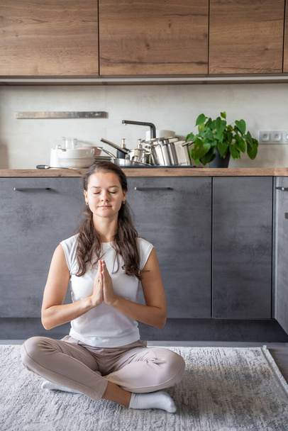 Young woman is sitting and doing meditation on background of dirty dishes in the kitchen. Slowdown, self-care, slow life concept. High quality photo - Photo, image