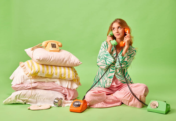 Wake up calls. Full length portrait of dissatisfied young woman in pajama talked to two retro phones isolated on pastel green background. Concept of wake up tome, healthy nap, work and rest, bedtime. - 写真・画像