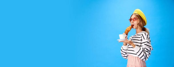 Schöne stylische junge Frau sieht in Baskenmütze mit leckerem Croissant und Tasse Kaffee auf blauem Hintergrund schockiert. Konzept von Essen, Küche, Mode, Stil. banner. Plakat mit Kopierraum für Text. - Foto, Bild