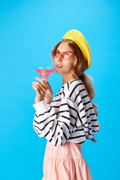 Portrét dámy oblečené v módním oblečení s růžovými slunečními brýlemi a žlutým baretem držet sklenici koktejlu na modrém pozadí. Koncept jídla a pití, alkohol, kuchyně, styl, dobrodružství. Reklama - Fotografie, Obrázek
