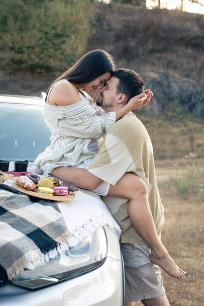Mooi jong paar genieten van picknick tijd op de zonsondergang in een weide, leunend op ouderwetse auto, man en vrouw genieten van elkaar. - Foto, afbeelding