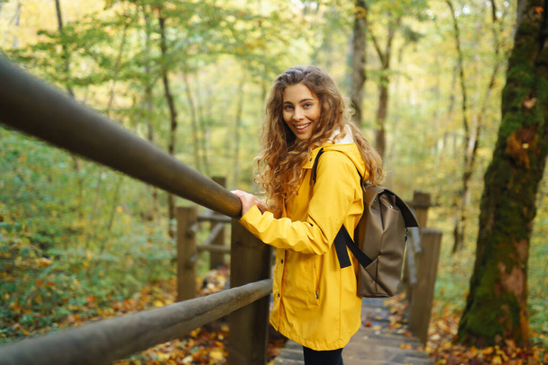 Active woman with backpack enjoys hiking on wooden stairs in autumn forest. A tourist female is engaged in mountaineering along a forest path, having fun, exploring nature. - Foto, immagini
