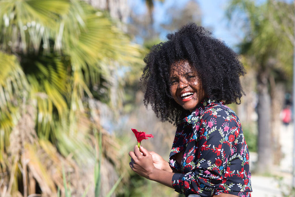 Jonge, mooie zwarte vrouw met afrohaar houdt een rode bloem in haar handen. De mooie vrouw is gelukkig en glimlachend - Foto, afbeelding