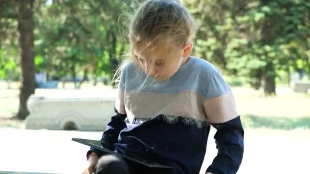 Klein meisje gebruikt een tablet tijdens het zitten in het park in de zomer, kijken naar het scherm. - Video