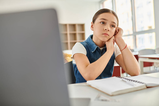 Ein junges Mädchen vertieft in Gedanken an ihrem Schreibtisch in einem lebhaften Klassenzimmer - Foto, Bild
