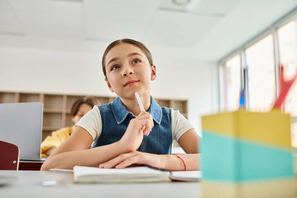 Молодая девушка, сидящая за столом, глубоко в мыслях, держит ручку, окруженную ярким классом - Фото, изображение