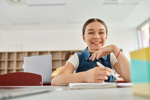 Ein fröhliches junges Mädchen sitzt an einem Schreibtisch und lächelt strahlend, während sie sich auf ihre Umgebung einlässt.. - Foto, Bild