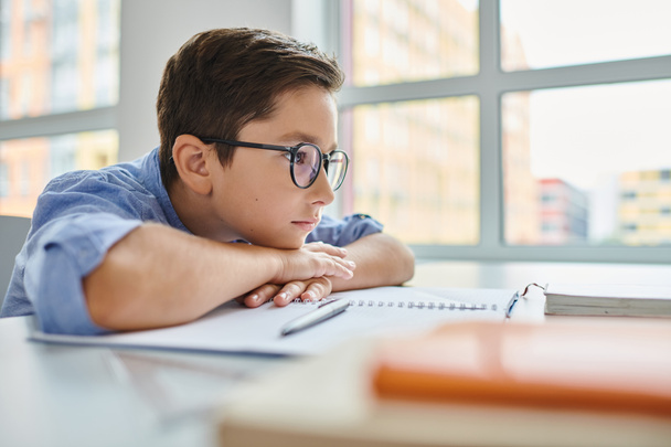 Ein fokussierter kleiner Junge mit Brille sitzt aufmerksam am Schreibtisch, vertieft in das Lernen oder die Arbeit an einer Aufgabe. - Foto, Bild