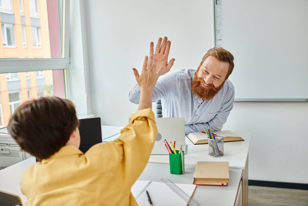 Ένα αγόρι με κίτρινη μπλούζα δίνει το "κόλλα πέντε" σε ένα δάσκαλο σε ένα φωτεινό, ζωντανό περιβάλλον στην τάξη. - Φωτογραφία, εικόνα