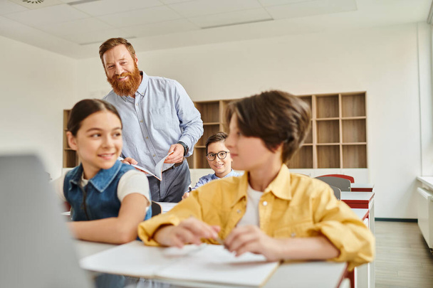 Μια ομάδα παιδιών κάθονται με προσοχή σε γραφεία σε μια φωτεινή τάξη, απορροφημένη από το μάθημα του υπολογιστή που διδάσκεται από τον αρσενικό δάσκαλό τους.. - Φωτογραφία, εικόνα