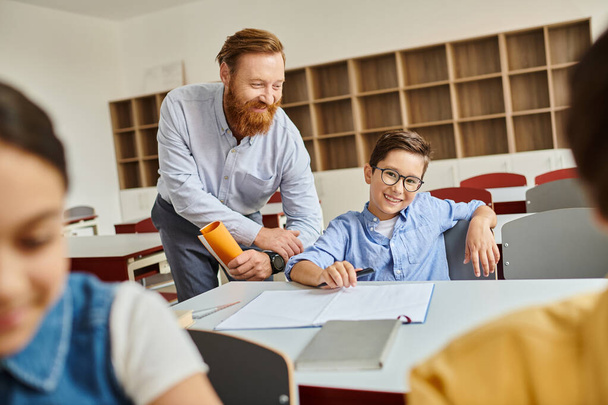 Ένας άνδρας στέκεται δίπλα σε ένα αγόρι σε μια πολύχρωμη τάξη, που ασχολείται με τη δυναμική μάθηση και τη διδασκαλία. - Φωτογραφία, εικόνα