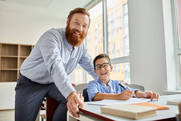 Een man die naast een jongen aan een bureau staat en bezig is met een leeractiviteit in een levendige klas. - Foto, afbeelding