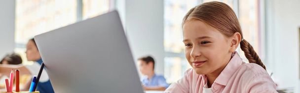 Egy fiatal lány, aki egy laptop előtt ül egy fényes osztályteremben, a virtuális világ felfedezésével foglalkozik. - Fotó, kép