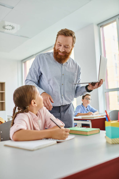 Мужчина стоит рядом с маленькой девочкой в ярком классе, участвуя в беседе один на один, в то время как остальные дети активно учатся вокруг них.. - Фото, изображение