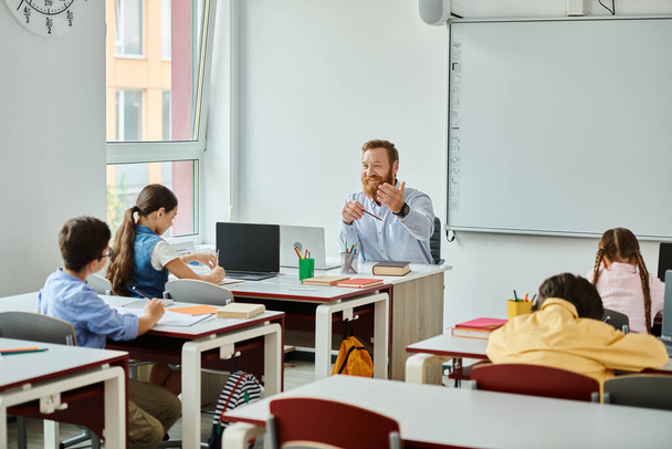 Un maestro masculino se sienta ante un grupo de estudiantes en un ambiente brillante y animado, participando en una instrucción interactiva. - Foto, imagen