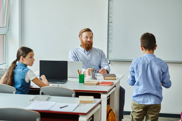 Un uomo si siede ad una scrivania di fronte ad un gruppo di bambini, coinvolgendoli in un ambiente vivace in classe. - Foto, immagini