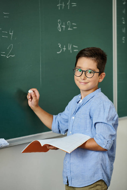 Een jonge jongen staat vol vertrouwen voor een schoolbord, bezig met het leren in een levendige klas. - Foto, afbeelding