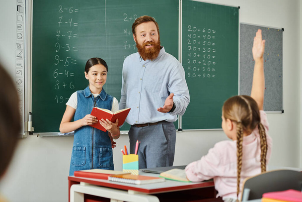 男は黒板の前で小さな女の子の隣に立って,活気に満ちた教室で教えています. - 写真・画像