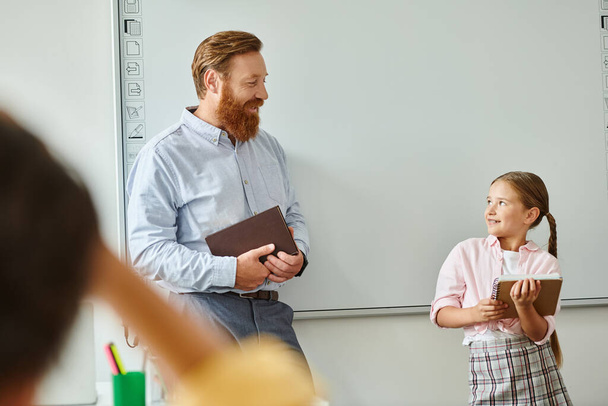 A man stands beside a little girl in front of a whiteboard in a bright classroom, engaging in interactive learning. - Photo, Image