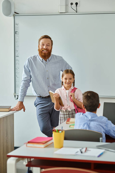 男はホワイトボードの前に立って,彼らが一緒に学ぶことに従事するように明るく活発な教室の設定で小さな女の子を指示します. - 写真・画像