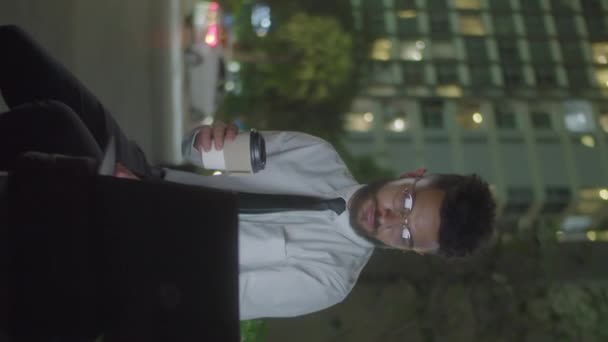 Tiro vertical del hombre de negocios sentado en el banco de la calle en la ciudad, sosteniendo para ir taza de café y usando el ordenador portátil mientras que trabaja en línea al aire libre en la noche - Imágenes, Vídeo