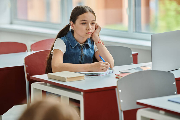 Una giovane ragazza si siede a una scrivania, con espressione del viso annoiata a scuola - Foto, immagini