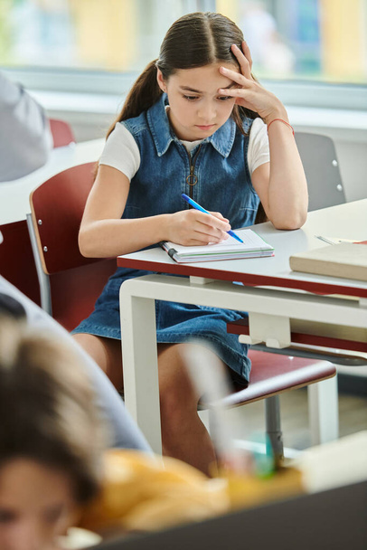Egy fiatal lány ül egy asztalnál egy jegyzetfüzettel és tollal, teljesen lefoglalja az írása.. - Fotó, kép