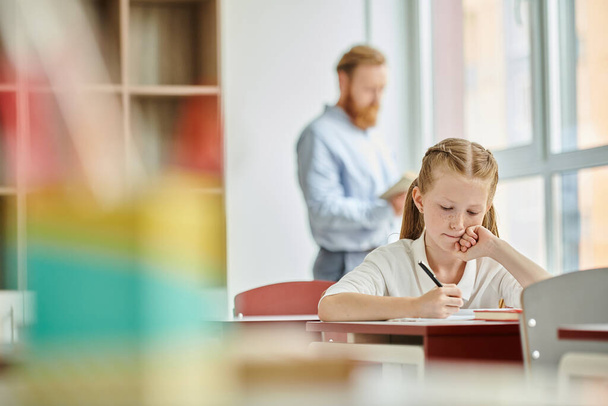 Una giovane ragazza si siede ad una scrivania, impegnata con uno studio in una classe luminosa e vivace mentre l'insegnante istruisce dietro di lei. - Foto, immagini