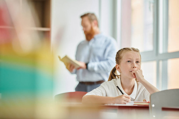 Une jeune fille s'assoit à une table tandis qu'un professeur d'homme se tient derrière elle, instruisant la classe - Photo, image