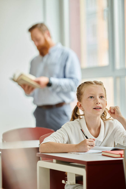 Ein kleines Mädchen sitzt aufmerksam an ihrem Schreibtisch und nimmt die Lektion eines Mannes in einem lebhaften und einladenden Klassenzimmer auf.. - Foto, Bild