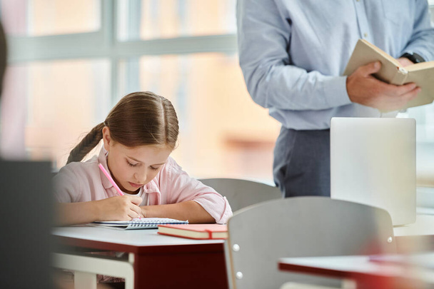 Een jong meisje zit aan haar bureau, verdiept in een boek en met behulp van een rekenmachine. Het klaslokaal is helder en levendig, met een leraar die haar instrueert - Foto, afbeelding