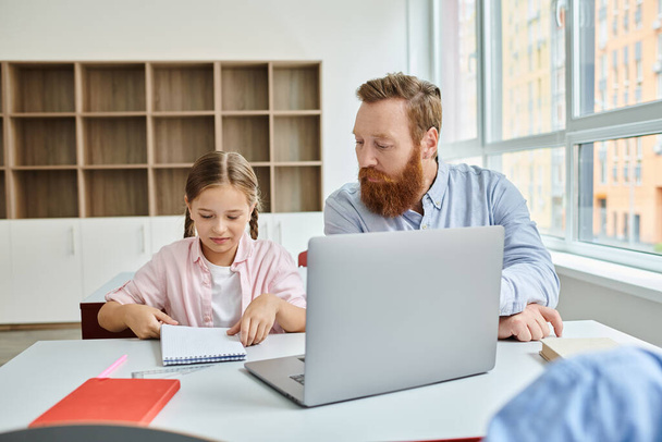 A man and a little girl sit attentively in front of a laptop, engaging in educational content during a lively classroom session. - Photo, Image