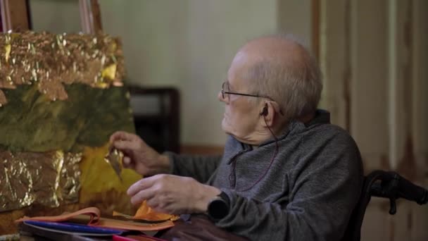 Ανώτερος καλλιτέχνης σε μια αναπηρική καρέκλα εφαρμόζει χρυσό στην ελαιογραφία σε καμβά - Πλάνα, βίντεο