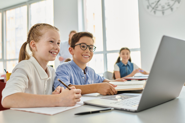 Eine Gruppe von Kindern sitzt an einem Tisch, konzentriert auf einem Laptop in einem hellen Klassenzimmer. - Foto, Bild