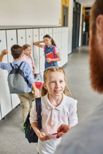 Çocuklar canlı bir koridordaki dolapları hevesle kontrol ediyorlar. Parlak bir sınıftaki erkek öğretmen rehberliğinde.. - Fotoğraf, Görsel