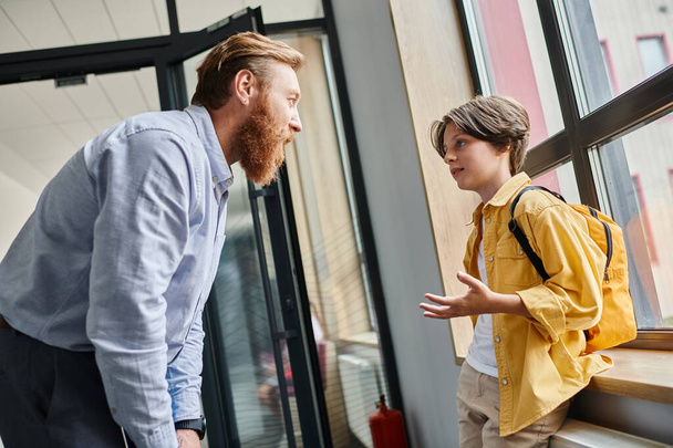 Een man staat naast een kleine jongen voor een raam, bezig met een bedachtzaam gesprek terwijl hij naar buiten kijkt. - Foto, afbeelding