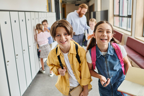 Un grupo diverso de niños camina por un pasillo de la escuela lleno de casilleros coloridos, charlando y riendo mientras se dirigen a su próxima clase. - Foto, imagen