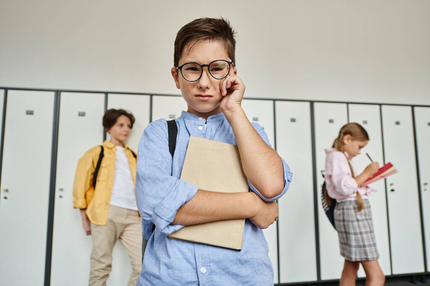 Ένα αγόρι με μπλε πουκάμισο στέκεται ανάμεσα σε σειρές ντουλαπιών στο διάδρομο του σχολείου.. - Φωτογραφία, εικόνα