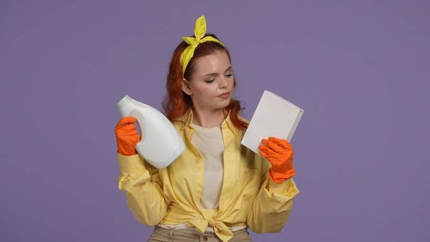 Everyday cleaning and housekeeping concept. Woman in casual clothing and rubber gloves holding box of washing powder and bottle, choosing between them. Isolated on purple background. - Foto, Imagen